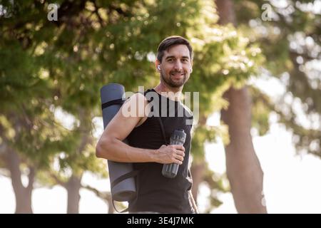 Smiling man wearing sporty top carries a yoga mat and water bottle while walking outdoors in a sunny park, showing a red birthmark on his arm, healthy Stock Photo