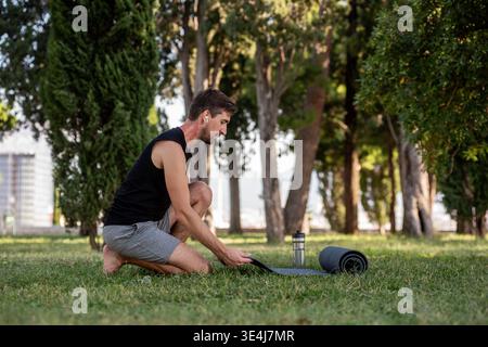 Fit man kneels on grass in a park, preparing his yoga mat and equipment for an outdoor practice. Red birthmark on his neck visible, focused on sport a Stock Photo