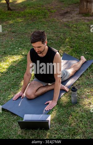 Focused man in sporty outfit practicing seated yoga pose on a mat in a sunny park, following an online class on laptop. Stock Photo