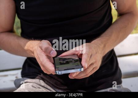 Close-up of a man in a black tank top holding and typing on a smartphone while sitting on a wooden bench outdoors, showing casual mobile technology us Stock Photo