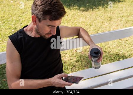 Young man in black sportwear sits on a white park bench after yoga, holding a shaker bottle and checking his smartphone. A red birthmark is visible on Stock Photo