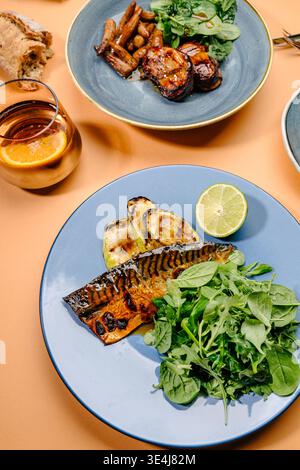 Overhead view of grilled mackerel with zucchini, spinach and arugula, served alongside bacon-wrapped pork medallions with baby corn on modern plates. Stock Photo
