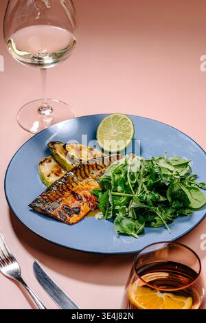 Grilled mackerel served with roasted zucchini, fresh spinach and arugula, and lime on a blue plate, styled on a pink table with wine glasses for a mod Stock Photo