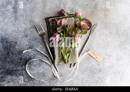 Elegant spring table setting featuring hellebore flowers on linen napkin, rustic silver cutlery and tag on gray stone background. Flat lay with soft n Stock Photo