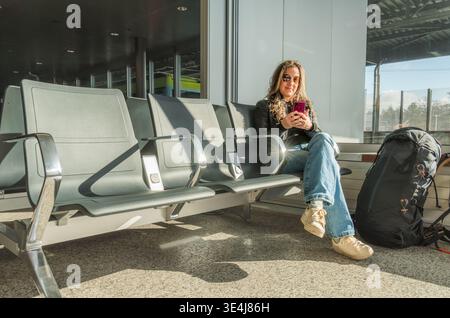 A woman in a casual outfit relaxes at a train station, using her smartphone. She is seated in a bright waiting area, with a large backpack beside her, Stock Photo