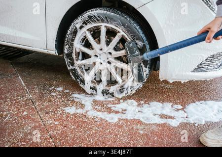 Close-up of a white car wheel being scrubbed with a brush and foamy soap on a wet driveway, showing hands cleaning the rim during an outdoor car wash. Stock Photo