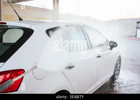 A white car being washed at a self-service car wash, sprayed with high-pressure water and foam. Car cleaning and detailing concept with wet pavement a Stock Photo