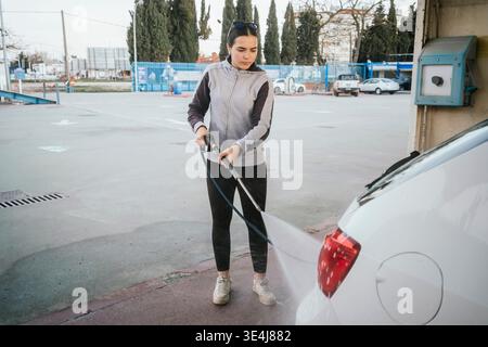 Focused teenager using a high pressure washer to clean a white car at an outdoor self service car wash bay, practicing responsibility and routine vehi Stock Photo