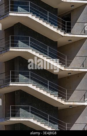 Close-up of a modern architecture building exterior featuring repeating concrete staircases, metal railings, and sharp geometric shadows, creating an Stock Photo