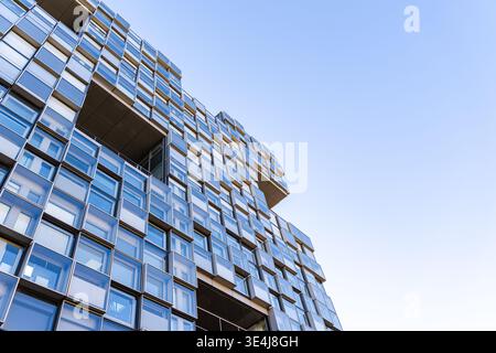 Low angle view of a modern glass building facade with stacked geometric windows and cantilevered terraces, reflecting blue sky. Contemporary urban arc Stock Photo