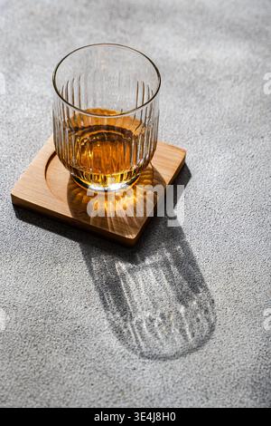 Sunlit glass of apple cider drink on a wooden coaster over gray concrete, casting a long sharp shadow. Minimal still life with warm amber tones. Stock Photo