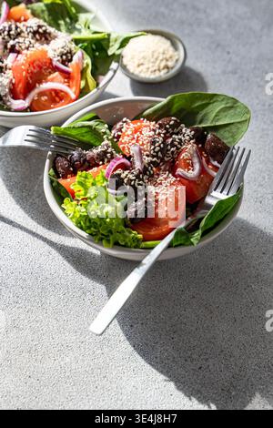 Fresh spinach salad bowl topped with sesame seeds, sliced tomato, black olives and red onion, served with a fork on a light stone background in natura Stock Photo