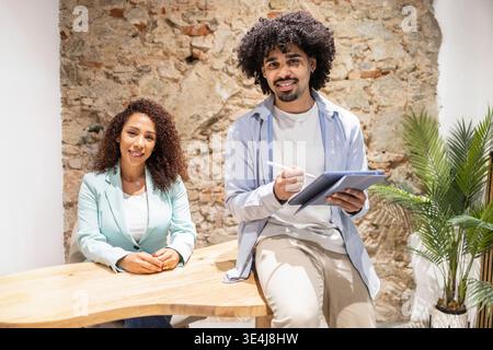 Two Latino coworkers in a bright coworking office, smiling and collaborating on a digital tablet beside a wooden desk, showing teamwork, creativity an Stock Photo