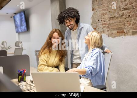 Smiling coworkers collaborate around a laptop in a modern coworking office, with a latin man and two colleagues sharing ideas, teamwork, and friendly Stock Photo