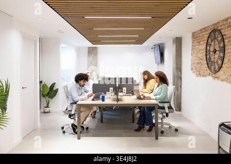 Diverse coworkers collaborate in a bright modern open office, seated at shared desks with laptops and screens, focused on teamwork, productivity, and Stock Photo