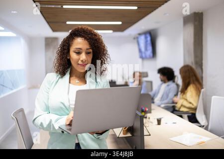 Confident professional woman working on a laptop in a modern coworking office, while a small team collaborates in the background, highlighting teamwor Stock Photo