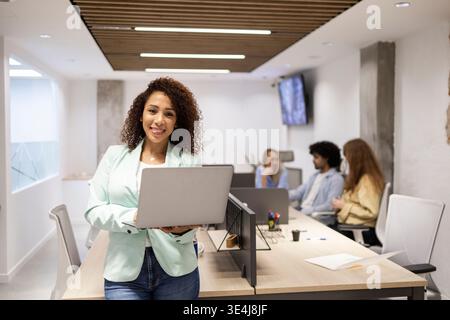 Confident young professional holding a laptop in a modern coworking office while teammates collaborate in the background, highlighting teamwork, produ Stock Photo