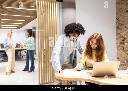 Latin man leaning over a table to help a coworker at a laptop in a modern coworking office, while teammates chat in the background. Teamwork, planning Stock Photo