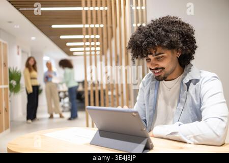 Smiling Latin man working on a tablet at a modern coworking office table while colleagues collaborate in the background, showing teamwork, productivit Stock Photo