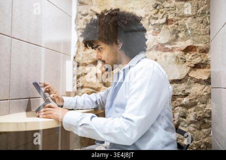 Side view of a latin man working in a modern office, using a tablet at a wooden desk. Focused professional in business shirt, against rustic stone wal Stock Photo