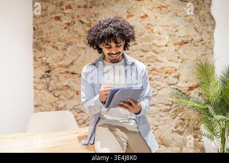 Smiling latin man in a modern office setting using a digital tablet, working remotely and reviewing notes. Natural light, casual style, productivity a Stock Photo