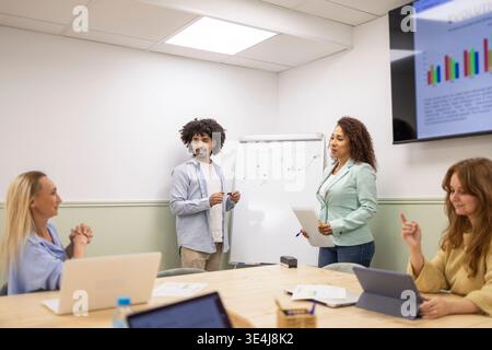 Diverse coworkers collaborate in a modern office meeting, reviewing charts on a screen and whiteboard while laptops are open, sharing ideas and planni Stock Photo