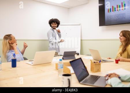 Diverse coworking team in modern office meeting room as a latin man leads a presentation at whiteboard, coworkers listening with laptops, planning str Stock Photo