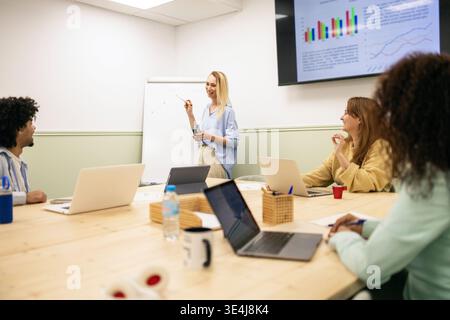 Diverse team collaborates in a modern coworking office during a strategy meeting, reviewing sales charts on a screen, sharing ideas around a table wit Stock Photo