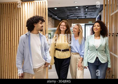 Diverse coworkers walk together in a modern coworking office hallway, smiling and talking during a team break, reflecting collaboration, startup cultu Stock Photo