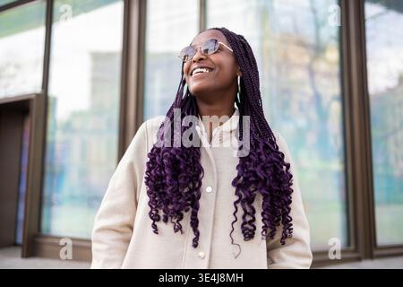 Smiling woman with sunglasses and purple braids enjoying a sunny day in the vibrant Gracia neighborhood, Barcelona. Reflects the urban lifestyle near Stock Photo
