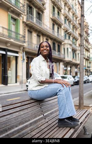 Young woman with braided hair sitting on a bench in the iconic Gracia neighborhood of Barcelona, Spain. The charming architecture enhances the urban l Stock Photo