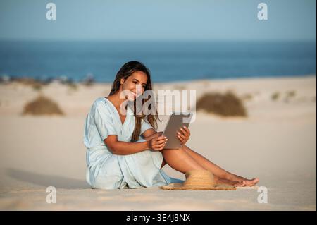 Young woman tourist sits on sandy dunes in Corralejo, using a tablet on a bright sunny day in Fuerteventura, Canary Islands, Spain, enjoying relaxed t Stock Photo