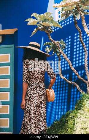Back view of a woman wearing a wide straw hat and leopard dress, holding a woven bag, walking past vivid blue walls and plants in a Moroccan street sc Stock Photo