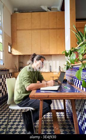 A man of Middle Eastern descent is seated at a wooden table in a kitchen. He is writing in a notebook while using a laptop, focusing on his tasks in d Stock Photo