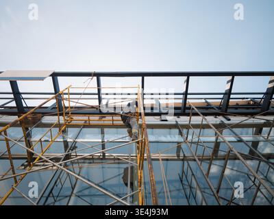 Low angle view of a construction worker on scaffolding installing glass panels on a modern building facade under a clear sky, highlighting safety, ren Stock Photo