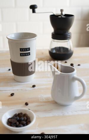 Minimal kitchen coffee scene with cold brew cup, manual grinder, small ceramic creamer pitcher, and scattered beans on a light wooden table, soft natu Stock Photo