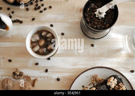 Top view of iced black coffee in a white ceramic mug, centered on a rustic table, with a modern black manual grinder packed with dark roasted beans in Stock Photo