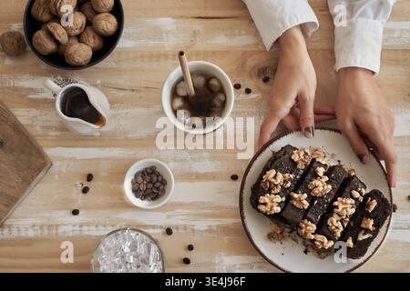 Top view of hands serving rich chocolate brownie slices topped with walnuts on a rustic wooden table, with iced coffee, cream, cocoa nibs, and whole w Stock Photo
