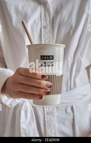 Close-up of a woman’s hands holding a white coffee mug labeled cold brew, with a straw, set against the soft texture of a white robe for a clean, cozy Stock Photo