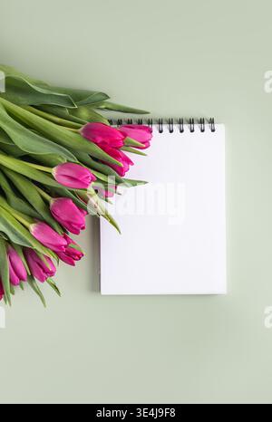 A vibrant pink tulip bouquet lies next to a blank spiral notebook on a soft green background, offering a fresh, serene scene perfect for creativity an Stock Photo