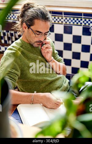 A man of Hispanic descent is sitting at a table in his kitchen. He is on the phone and writing notes in a notebook. Green plants surround him, adding Stock Photo