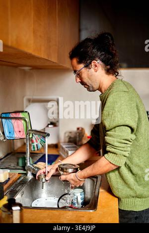 A man of Hispanic descent is working from home in his kitchen. He is washing dishes and managing chores while possibly preparing for remote work tasks Stock Photo