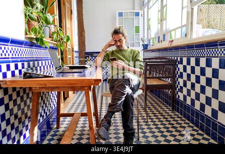 A man sits in a room with blue tiles, working from home. He focuses on his smartphone while his laptop is open on the table. Bright light comes throug Stock Photo
