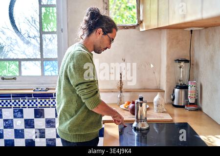 A man with curly hair and glasses is making coffee in a kitchen. He stands by the counter, focused on a stove. Plants and fruits are on the counter, s Stock Photo