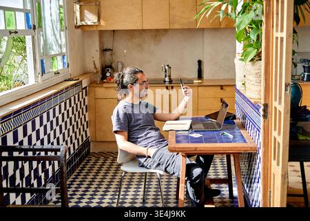 A man of Hispanic descent sits at a table in a kitchen. He uses a laptop and checks his phone. Sunlight enters through the window. The kitchen has til Stock Photo
