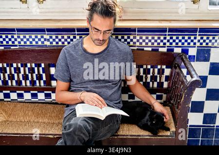 A man of Hispanic descent sits on a bench at home reading a book. A black cat rests next to him. The room has blue and white tiles. Natural light fill Stock Photo