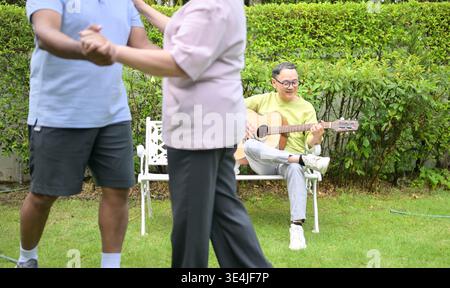 Group of asian seniors singing and dancing while their friend plays guitar in a garden, enjoying friendship and outdoor leisure activity || Model released Stock Photo