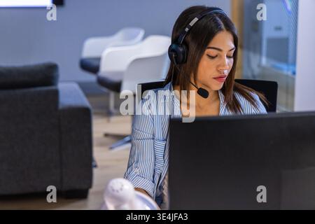 Asian woman sitting at desk wearing buttonup shirt, headset, using laptop with device in office. Callcenter, lounge, professional, headphones, microphone, workspace, openplan || Model released Stock Photo