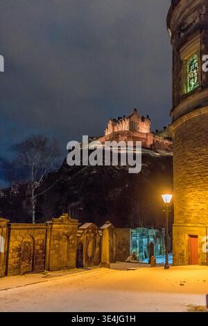 A moody nighttime view of Edinburgh Castle perched on a rocky crag, photographed from St John's churchyard with warm streetlamp light, snow-covered gr Stock Photo