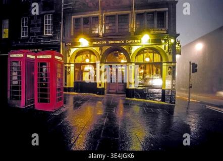 Warm-lit Deacon Brodie's Tavern glows on Edinburgh's Royal Mile at night, framed by classic red telephone boxes, wet cobblestones and moody fog reflec Stock Photo